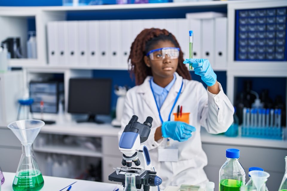 Technicienne de laboratoire en blouse et lunettes de protection, tenant un tube a l'examen devant un microscope